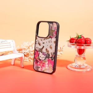 以下が含まれることがあります： 黒い携帯電話ケース。ハローキティのグラフィック、ピンクの花、落書き風のテキストをあしらったカラフルなデザイン。ケースは黒い縁と透明な背面を備えています。ヒョウ柄の星も特徴です。