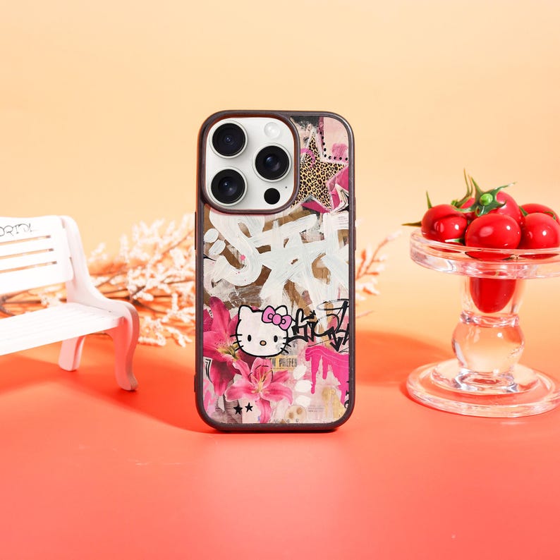 May include: A phone case featuring a Hello Kitty design with a graffiti-style background. The case has a black border and a colorful design with pink flowers, a leopard print star, and white graffiti-style text. The Hello Kitty character is in the lower center.