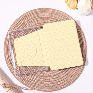 Peut inclure: Un étui jaune à pois, ouvert pour révéler une fente pour carte. Un étui en plastique transparent est à côté. Les articles sont sur un dessous de plat rond et tissé.