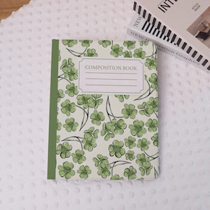 Pode incluir: Um caderno de composição com uma capa creme com um padrão de trevos verdes de quatro folhas. O livro tem uma lombada verde e as palavras "COMPOSITION BOOK" estão impressas na capa. Há também um rótulo branco.