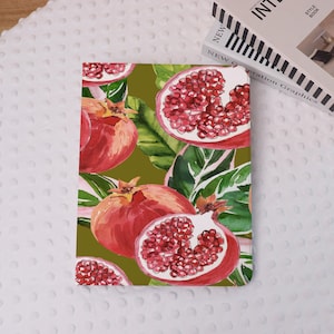 Op de afbeelding: Een notitieboek met een aquarelontwerp van granaatappels en groene bladeren op een donker olijfgroene achtergrond. De omslag toont hele en gesneden granaatappels met zichtbare zaden. Het notitieboek ligt op een witte, getextureerde ondergrond.