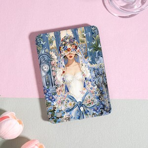May include: A rectangular decorative plaque featuring a detailed illustration of a person wearing a white dress and a floral veil over their eyes. The scene includes a blue grandfather clock, cascading blue flowers, and butterflies in a room.