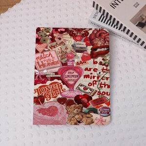May include: A notebook with a collage design featuring hearts, text, and a perfume bottle. The color palette includes shades of red, pink, and white. The text includes "Self Love Potion", "Match Made in Heaven", and "Lover Girls". A book is visible in the background.