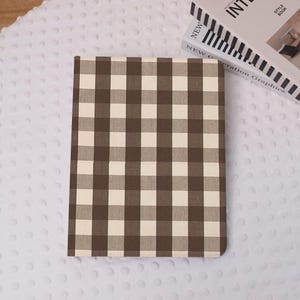 Op de afbeelding: Een bruin en crèmekleurig geruit notitieboek of dagboek. De omslag heeft een klassiek ruitpatroon. Het notitieboek is rechthoekig en gesloten. Een witte, getextureerde achtergrond is zichtbaar, samen met een boek.