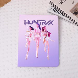 Può includere: Una copertina di taccuino viola chiaro con la scritta "HUNTRAX" e tre figure animate in abiti bianchi e dorati. Le figure sono in posa dinamica su uno sfondo sfumato. Un libro è visibile sullo sfondo.