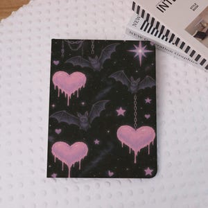 Pode incluir: Caderno preto com um padrão de morcegos, corações rosa pingando e estrelas em um fundo estrelado. O caderno tem um design de corrente e está ao lado de dois livros.