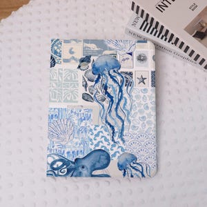 Peut inclure: Une housse de tablette à motifs bleus et blancs avec un collage d'illustrations de la vie marine. Le motif comprend des méduses, des coquillages et une pieuvre. L'étui est rectangulaire et a une surface lisse.
