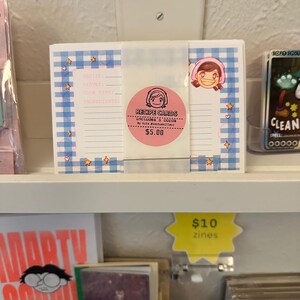 May include: A stack of white recipe cards featuring a blue gingham border with small orange star and pink heart accents. A pink circular sticker in the center displays a cartoon face and the text RECIPE CARDS INCLUDES 8 CARDS By kris pootuskillerz.