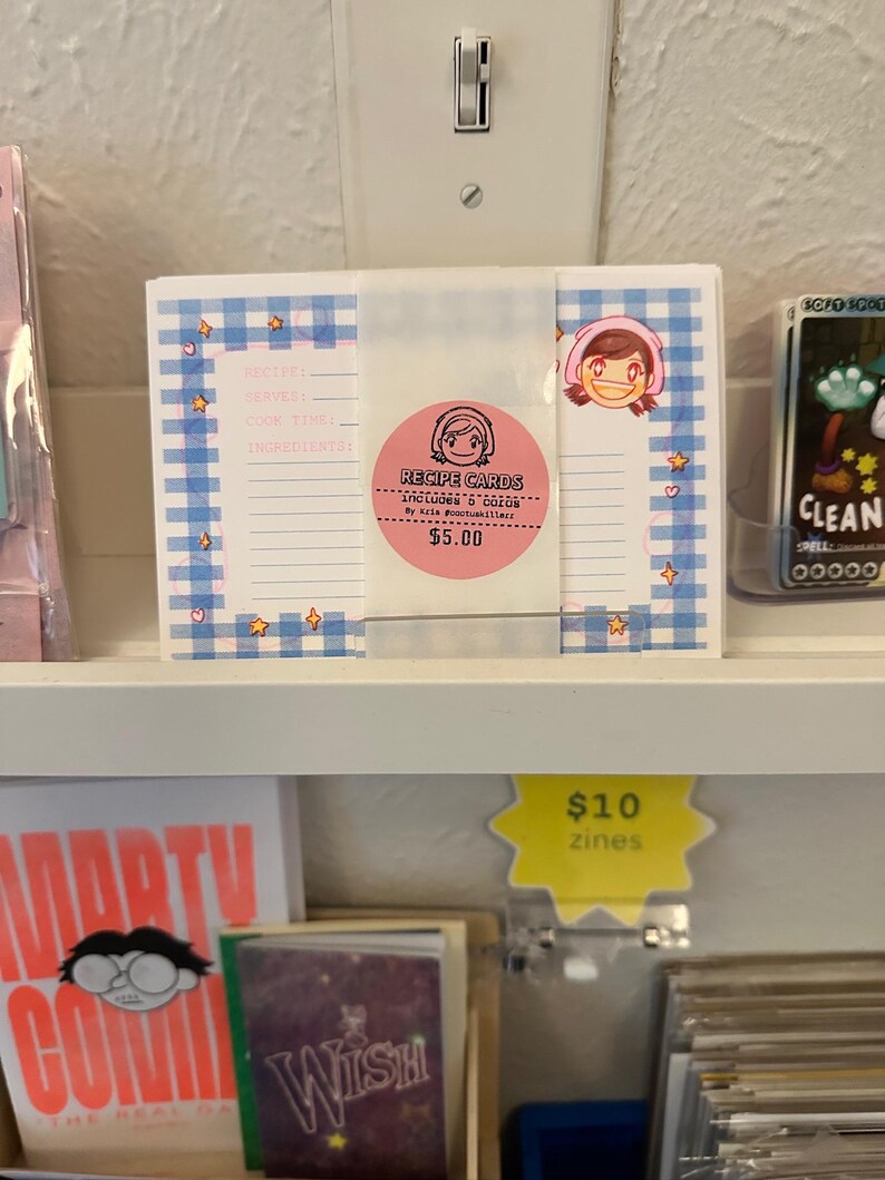 May include: A stack of white recipe cards featuring a blue gingham border with small orange star and pink heart accents. A pink circular sticker in the center displays a cartoon face and the text RECIPE CARDS INCLUDES 8 CARDS By kris pootuskillerz.
