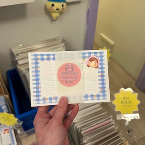 May include: A rectangular set of recipe cards featuring a blue and white gingham border with small star and heart illustrations. A central pink circular sticker displays the text "RECIPE CARDS includes 5 cards" and a cartoon character illustration.