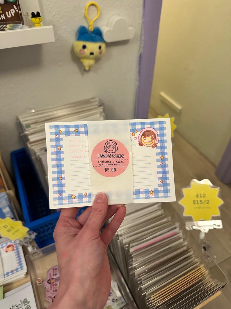 May include: A rectangular set of recipe cards featuring a blue and white gingham border with small star and heart illustrations. A central pink circular sticker displays the text "RECIPE CARDS includes 5 cards" and a cartoon character illustration.