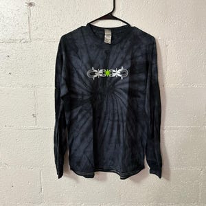 May include: Black and gray tie-dye long-sleeve shirt with a white and green graphic design. The shirt is hanging on a black hanger against a white brick wall.