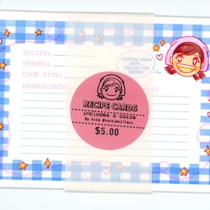 May include: A rectangular recipe card featuring a blue and white gingham border with small orange stars and pink hearts. The card includes printed lines for recipe details and a cartoon character. A pink circular sticker reads RECIPE CARDS.