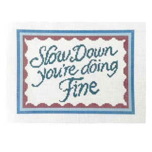 Op de afbeelding: Naaldkunst canvas met de tekst "Slow Down You're Doing Fine" in turquoise schrift. De tekst is gecentreerd op een witte achtergrond, omlijst door een geschulpte rand in rood en een blauwe rand.