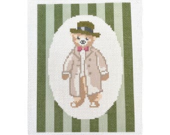 May include: A needlepoint canvas featuring a brown teddy bear wearing a green hat, a white coat, and a pink bow tie. The bear is standing in a white circle on a green and white striped background.