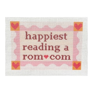 May include: A needlepoint design with the text "happiest reading a rom*com" in brown letters. The text is inside a pink wavy border, with a pink heart in the word "rom*com". The corners have orange squares with pink hearts.