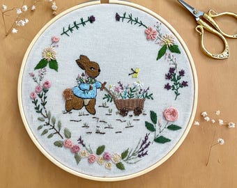 May include: Embroidered hoop art featuring a brown rabbit pushing a wheelbarrow filled with flowers. The hoop is surrounded by a wreath of green leaves and pink, white, and purple flowers.