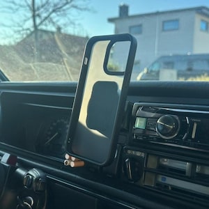 Peut inclure: Un étui de téléphone transparent avec un bord noir est monté sur le tableau de bord d'une voiture. L'étui est vide et l'intérieur de la voiture est visible. Des cigarettes sont placées dans un support sous l'étui.