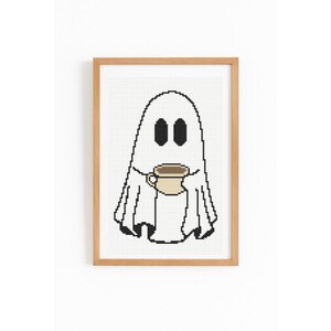 May include: A framed cross-stitch art piece featuring a white ghost holding a coffee mug. The ghost has black oval eyes and a black outline. The coffee mug is beige with a brown coffee fill. The frame is a light brown color.