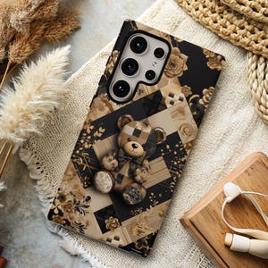 Könnte beinhalten: Eine schwarze Handyhülle mit einem gold-braunen geometrischen Muster und einem Teddybär-Design. Die Handyhülle hat einen Ausschnitt für das Kameraobjektiv.