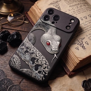 Puede incluir: Una funda de smartphone negra con una serpiente plateada en relieve, detallada con ojos rojos llamativos y patrones de escamas complejos. El diseño incluye elementos decorativos en espiral que aportan una apariencia texturizada al dispositivo.