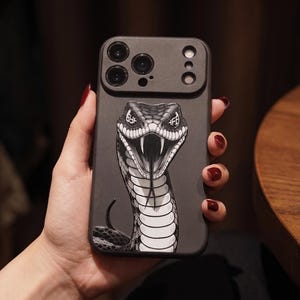 May include: A matte black smartphone case featuring a high-contrast, black and white graphic illustration of a snake with its mouth open, showing fangs and a forked tongue. The snake's eyes contain the white text characters 6YF and HFB.