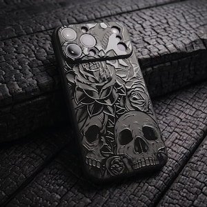 Op de afbeelding: Een zwarte smartphonehoes met een gedetailleerd monochroom gegraveerd ontwerp van meerdere menselijke schedels en bloeiende rozen. De hoes bevat een verhoogde rechthoekige uitsparing voor de cameramodule met ronde openingen.
