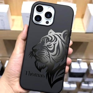 以下が含まれることがあります： 黒色のスマートフォンケース。 灰色の濃淡で描かれた詳細な虎のデザインが特徴です。「Thomas」という名前が刻まれています。 ケースは滑らかな仕上がりで、スマートフォン向けに設計されています。