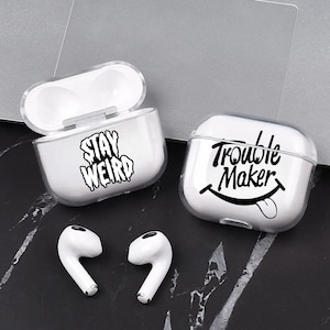 Puede incluir: Fundas blancas transparentes para AirPod con texto negro que dice "Stay Weird" y "Trouble Maker" con una cara sonriente. Dos Airpods blancos están al lado de las fundas. Las fundas están sobre una superficie oscura.