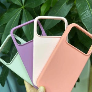 Pu&ograve; includere: Quattro custodie per telefono colorate nei toni del bianco, lavanda, rosa chiaro e corallo. Le custodie sono realizzate in materiale liscio e hanno una forma rettangolare con un ritaglio per la fotocamera. Lo sfondo presenta foglie verdi.