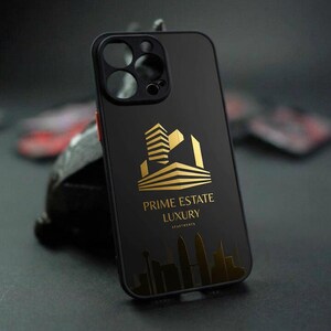 Pu&ograve; includere: Custodia per telefono nera con logo dorato "Prime Estate Luxury Apartments". Il logo presenta un design stilizzato di un edificio. La custodia ha un design elegante e moderno.