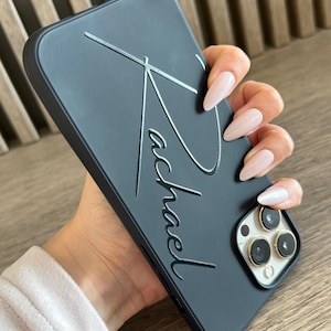 Puede incluir: Carcasa de teléfono negra con una firma en relieve plateada que dice "Michael".