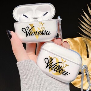 Può includere: Due custodie trasparenti in plastica per Airpods con un design floreale dorato e nomi personalizzati. La custodia a sinistra dice "Vanessa" e la custodia a destra dice "Willow".