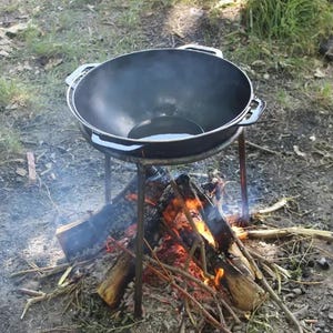 Puede incluir: Una olla de hierro fundido negra con un asa a cada lado se asienta sobre un trípode sobre una pequeña fogata. La olla está llena de aceite y está humeando.