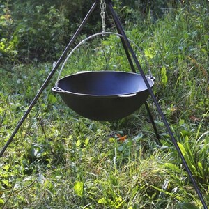 Puede incluir: Un caldero de hierro fundido negro colgado de un trípode sobre hierba verde. El caldero está suspendido por una cadena y un anillo de metal.