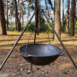 May include: A black cast iron cauldron suspended from a metal tripod with a chain. The cauldron has a rounded base and a handle. The tripod is set up outdoors in a wooded area with tall trees and dry grass.
