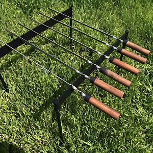 Op de afbeelding: Een zwarte metalen standaard met zes lange metalen spiesjes met houten handvatten. De standaard staat op een bed van groen gras.