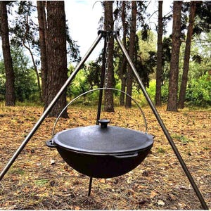 May include: A black cast iron cauldron with a lid, suspended from a metal tripod with a chain. The cauldron has a rounded shape and is designed for outdoor cooking. The tripod is made of metal and is set up in a forest setting.