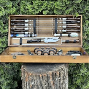 May include: A wooden box with a set of grilling tools. The box contains skewers, a spatula, tongs, a fork, and a small container of grilling sauce. The box is open and the tools are visible.