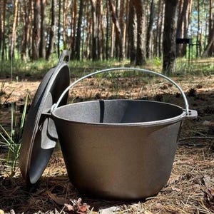 Op de afbeelding: Een donkergrijze gietijzeren Dutch oven met een deksel en een metalen handvat. Het deksel staat iets open en onthult de binnenkant. De Dutch oven staat op de grond in een bosrijke omgeving.
