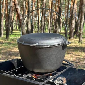 Puede incluir: Una olla de hierro fundido negra con tapa y asa está sentada sobre una rejilla de metal sobre un fuego. La olla está en un entorno boscoso.