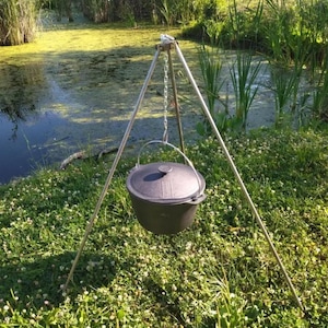 May include: A black cast iron pot hanging from a tripod stand over a green pond. The stand is made of metal and has three legs.