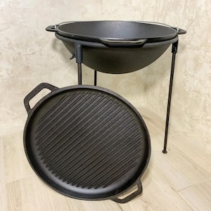 以下が含まれることがあります： 黒い鋳鉄製のダッチオーブンと、グリルパンの蓋と黒い金属製のスタンド。ダッチオーブンはスタンドの上にあり、蓋は横に置かれています。