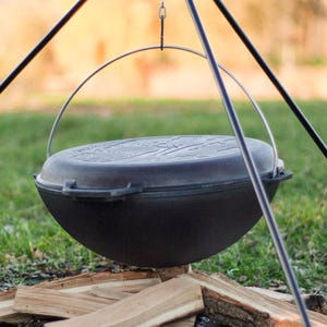 May include: A black cast iron pot with a lid hanging over a campfire on a tripod stand.
