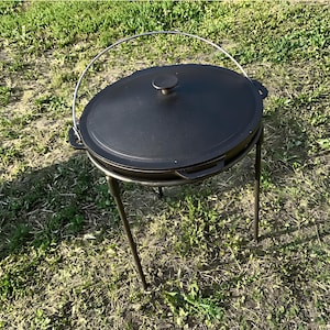 May include: Black cast iron Dutch oven with a lid and a metal handle. The oven is sitting on a black metal stand with three legs. The oven is on a bed of green grass.