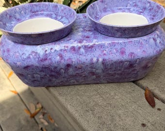 May include: A ceramic pet bowl with a lavender and white speckled design. The bowl has two circular openings on top and a rectangular base. The interior of the bowls are white. The bowl is sitting on a wooden deck.