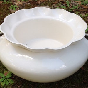 May include: A large, off-white ceramic planter with a scalloped rim. The planter has a wide, rounded base and two small handles. The design is simple and elegant, suitable for various plants and garden settings.