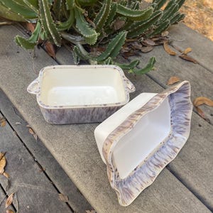 May include: Two rectangular ceramic baking dishes. The dishes are white inside with a speckled purple and brown exterior. One dish has handles on the sides. The other dish is tilted on its side. The dishes are on a wooden surface.
