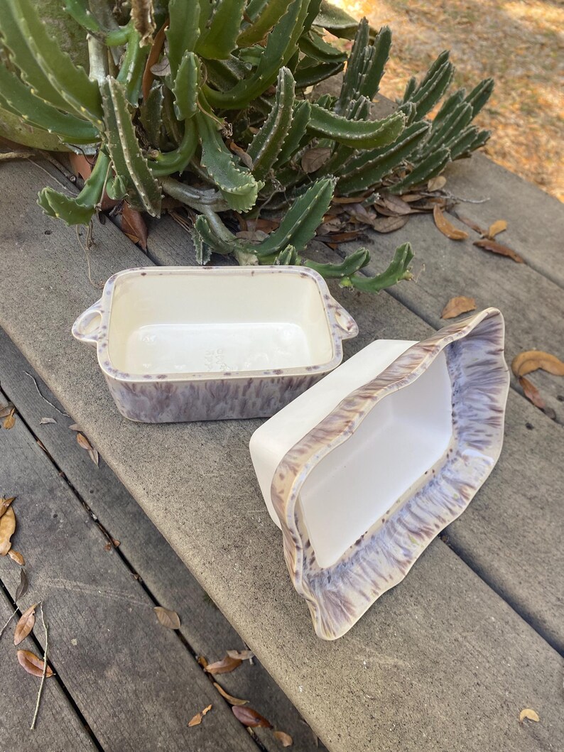 May include: Two rectangular ceramic baking dishes. The dishes are white inside with a speckled purple and brown exterior. One dish has handles on the sides. The other dish is tilted on its side. The dishes are on a wooden surface.