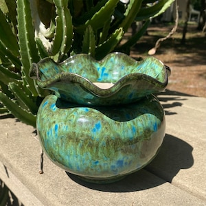 May include: A two-piece ceramic planter with a glossy finish. The bottom pot is round, and the top is a ruffled bowl shape. The planter is glazed in shades of green and blue, with dark brown accents. The planter is sitting on a concrete surface.